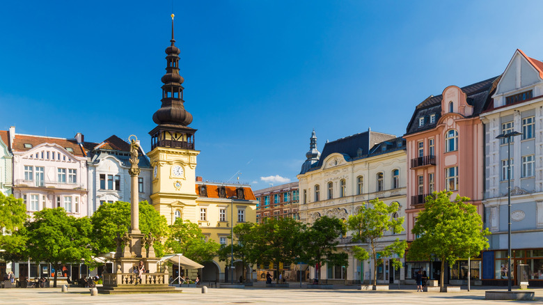 A town square with pastel-colored buildings and leafy trees, under a blue sky