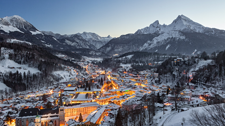 A small, lit village in the winter with large snow-covered mountains in the background