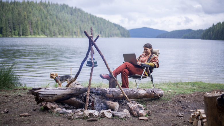Young woman having her morning coffee and using laptop for working remotely while on a caYoung woman having her morning coffee and using laptop for working remotely while on a camping vacation