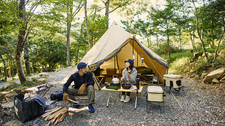 man prepares campfire while woman watches camping in the woods