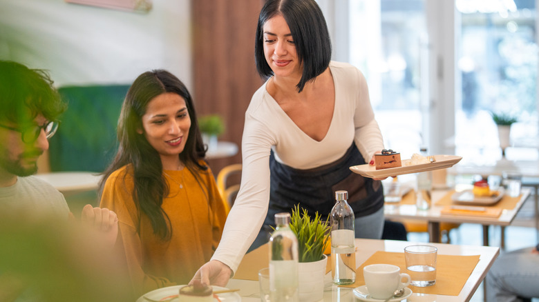 Waitress in a white long-sleeve top serving plates to a seated customer at a bright, contemporary restaurant.