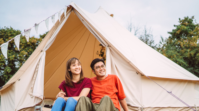 Young smiling Asian couple sitting in front of a setup glamping tent