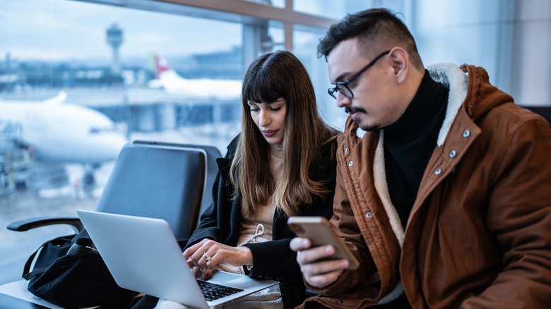 A man and woman use a mobile phone and laptop in the airport.