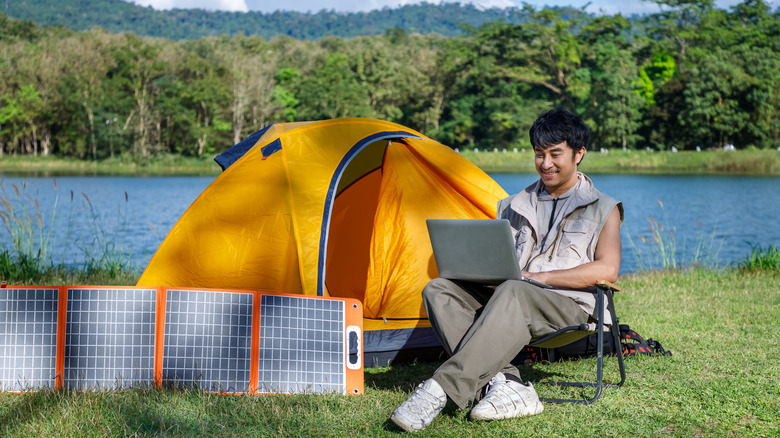 A young man sits with a computer outside a tent next to flexible solar panels.
