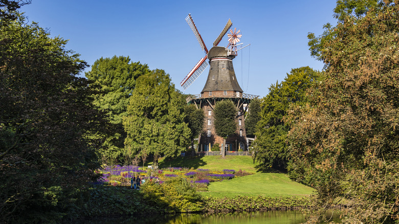 An old wooden windmill surrounded by trees and greenery in front of a pond