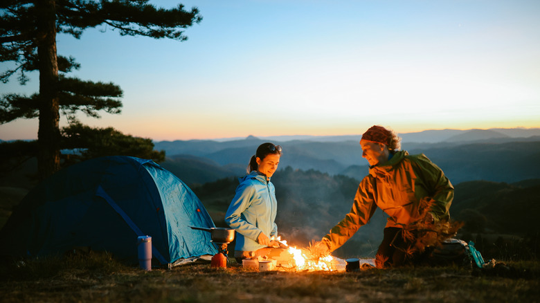 A mother and daughter camping outdoors with a tent around a campfire.