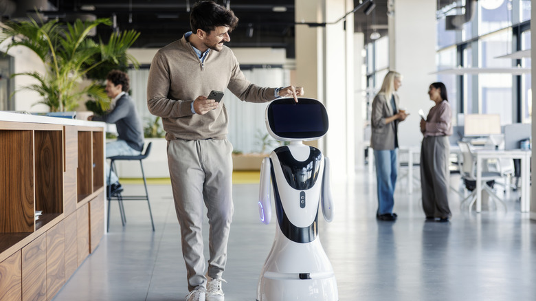 A young adult man using a smartphone and touching a robot's screen in a contemporary office space.