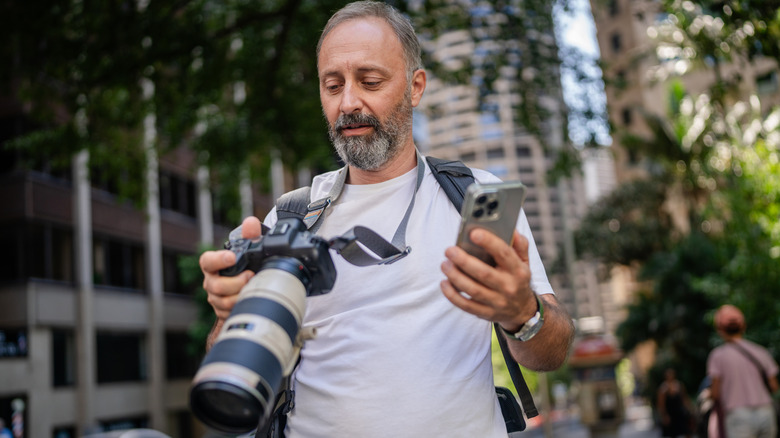 A man stands on a busy street in Sydney, Australia, adjusting his camera while holding a smartphone.