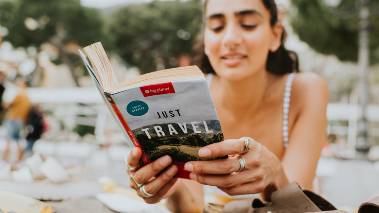 A beautiful Indian woman lies on her stomach on a busy beach reading a travel guide book.