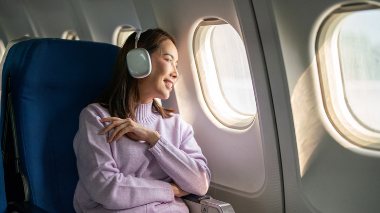 A passenger woman on a flight is seen relaxing with wireless headphones, enjoying the view outside the window as she travels to her destination