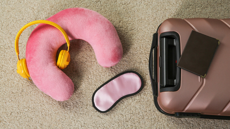 Pink travel pillow with headphones, suitcase, passport and sleep mask on beige rug, flat lay