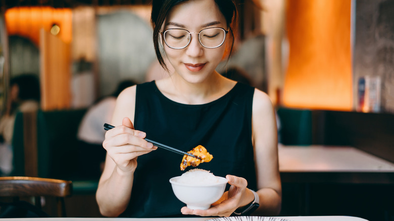 A woman wearing glasses and black shirt holds a dumpling with a pair of black chopsticks while holding it over a bowl of rice