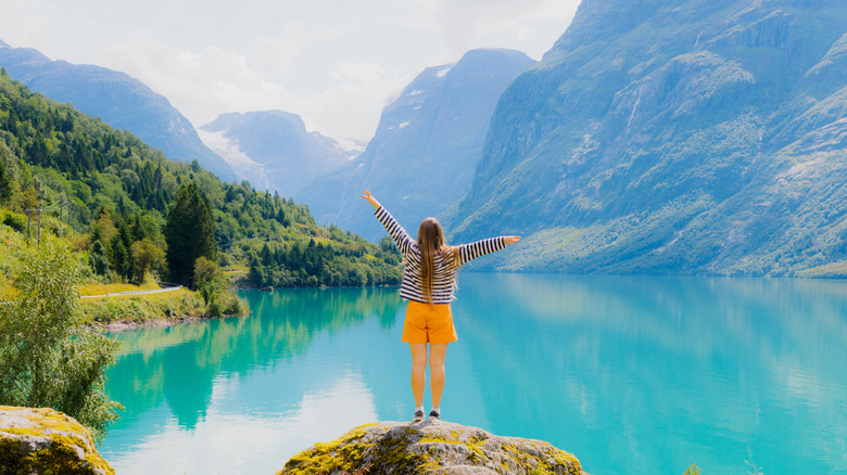 Happy female traveler in orange shorts standing on rock and facing a scenic viewpoint of glacial mountain Lake Lovatnet in Western Norway, Scandinavia