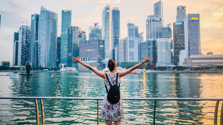 Young traveler woman with her arms outstretched watching sunset above the skyline in Singapore bay area.