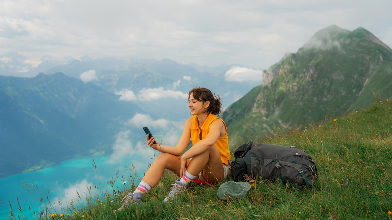 Woman sitting on the meadow in Swiss Alps overlooking Interlaken and using smartphone during the hike in summer