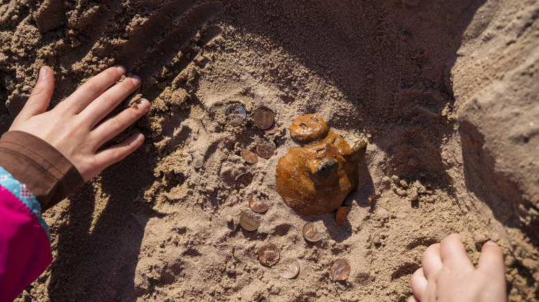 Childs hand digging a whole with a buried piggy bank and coins in it