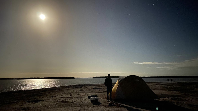 A man stands by a tent on the water's shore