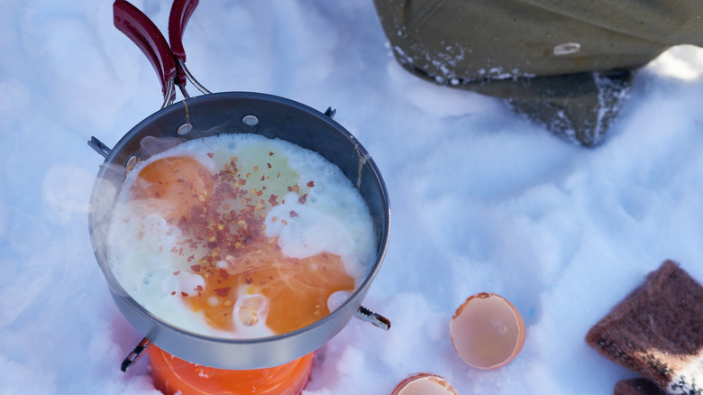 Cooking eggs in metal pan on portable stove outdoors in snow with cracked eggshells and bread nearby,