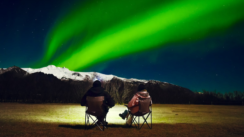 Romantic Couple in black and bright outfit sit in foldable camping chairs watch northern lights show