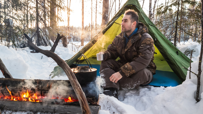 Man sat before a campfire against a wintery background.