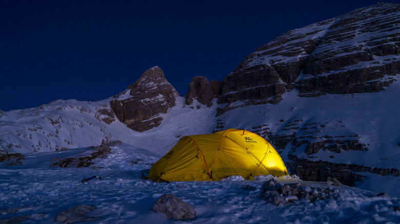 A glowing yellow tent rests on a snowy mountain slope under a dramatic alpine sky during sunrise. Rugged peaks surround the serene campsite.