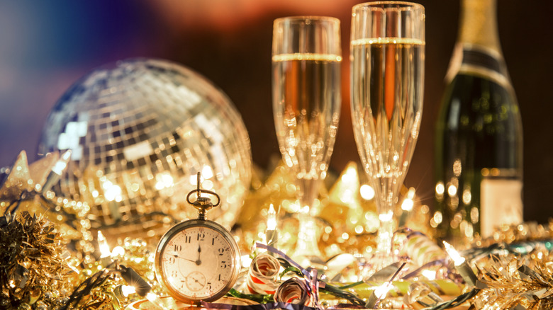 A disco ball, two glasses of champagne, a stopwatch counting down to midnight, a bottle of champagne, and a pile of gold and sparkly tinsel for New Year's Eve