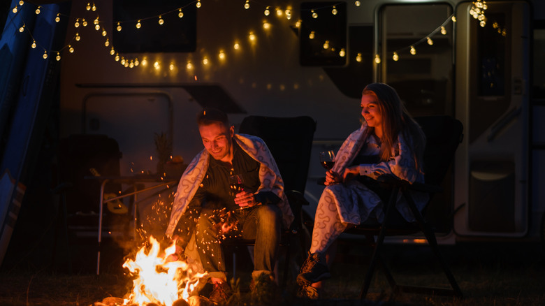 A man and woman sitting by a fire with string lights above them.