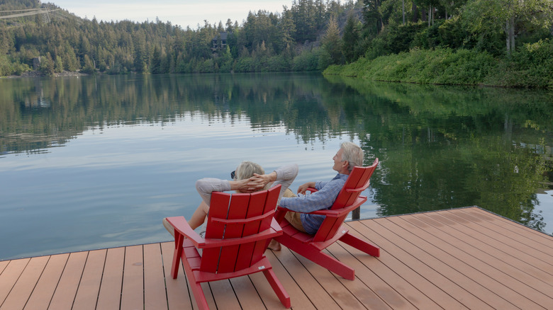 Senior couple sit in red chairs on a dock overlooking a peaceful lake rimmed with green pine trees