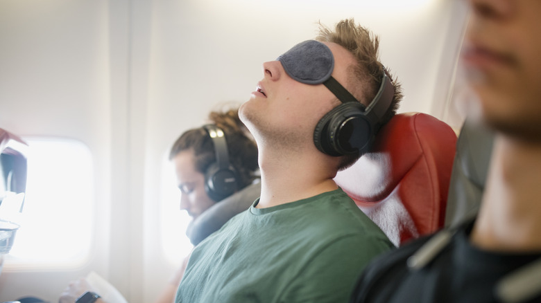 A young man asleep on a flight wearing wireless headphones and an eye mask.