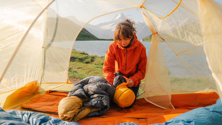 A camper packing up a sleeping bag and air mattress in a tent next to a lake and mountains.
