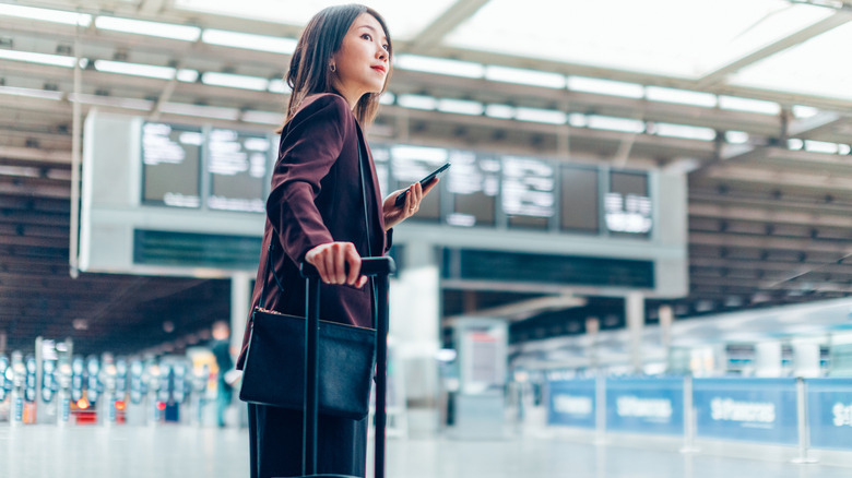 A woman holding a phone and suitcase at an airport departure area.