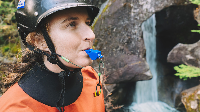 A helmeted woman near a waterfall clutches a whistle in her teeth