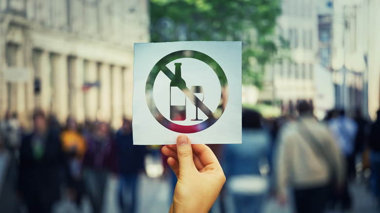 A hand holding a paper sheet with no alcohol sign over a crowded street background.