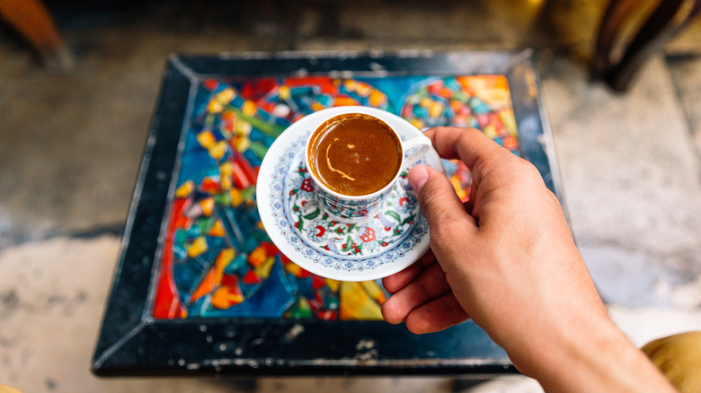A hand holds a saucer and cup full of Turkish coffee over a colorful table