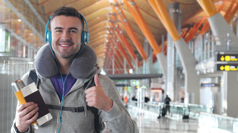 A male traveler giving a thumbs up wearing headphones and a neck pillow while holding his passport