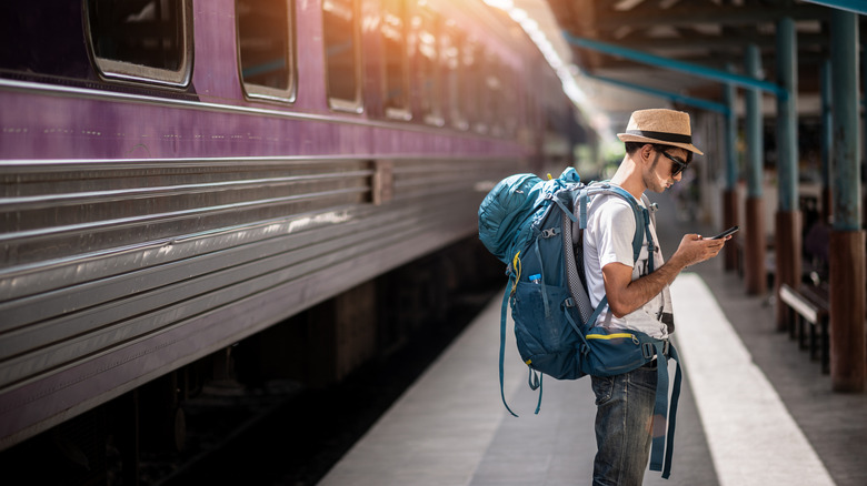 young male backpacker stands on train platform looking at cell phone