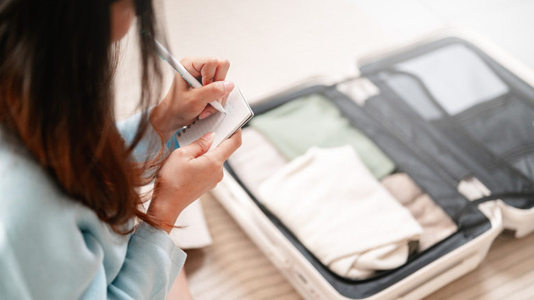 an open suitcase with clothes inside while a person writing on a piece of paper