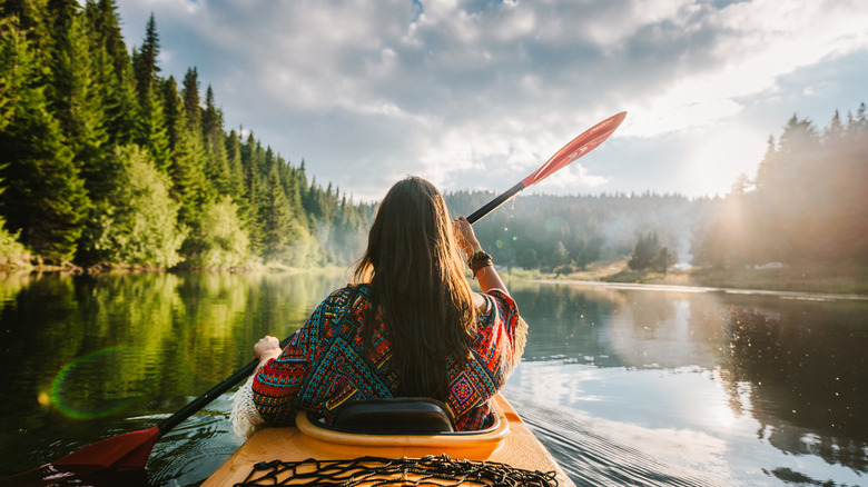 A brunette woman wearing a sweater paddles a yellow kayak on a misty, tree-lined lake