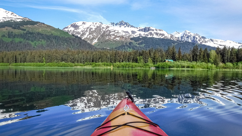 The front of a red kayak on a pristine lake framed by forest and snowy peaks near Seward, Alaska