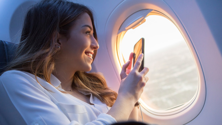 A smiling woman with long hair taking a photo while looking out of an airplane window