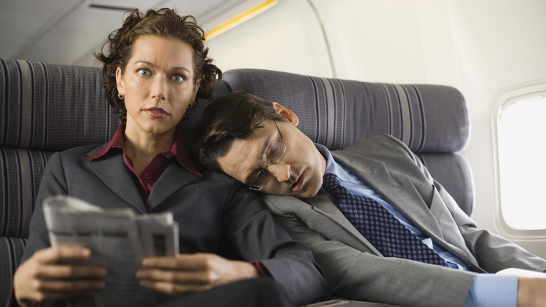 A business woman sitting in her airplane seat looking startled with a business man sleeping on her shoulder