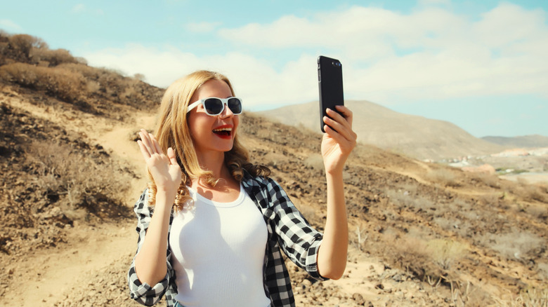 A cheerful woman waving her hand on a video call while hiking