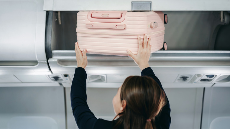 Female traveler stowing pink luggage overhead bin on passenger airplane preparing for flight journey inside modern cabin interior space