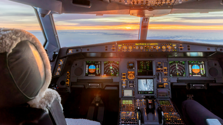 Interior view of a commercial jet cockpit during flight at high altitude, with avionics and navigation displays lit up as the sun rises on the horizon.