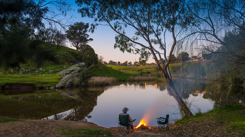 a man sits in front of a camp fire beside a billabong in Australia