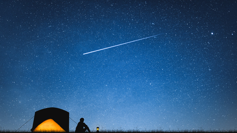 A camper sitting next to a tent gazing at a shooting star in the dark blue sky.