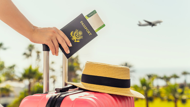 A traveler holding a passport and handle of a pink suitcase with a hat resting on the suitcase and a plane flying in the background.