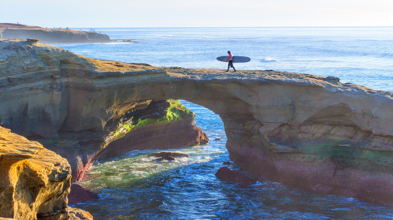 Surfer carrying board over natural arch near San Diego, California, on sunny day