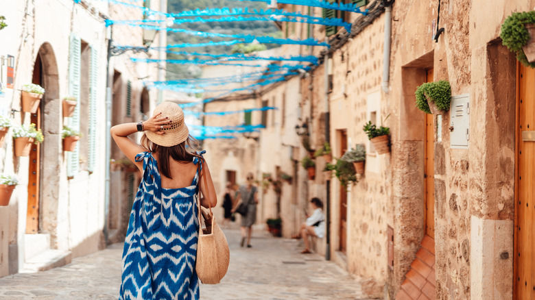 A rearview of a woman wearing a blue dress and hat walking through cobblestone streets