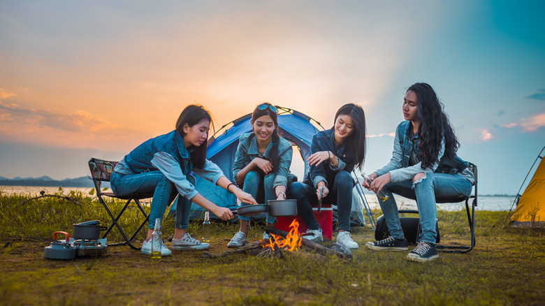 Four women camping and cooking over a fire in front of a tent.